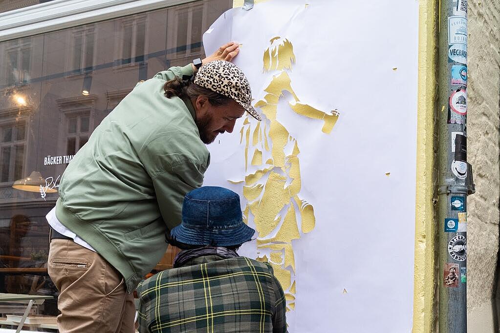 Zwei Personen befestigen eine Papierschablone an der Außenwand einer Bäckerei in der Flensburger Innenstadt.