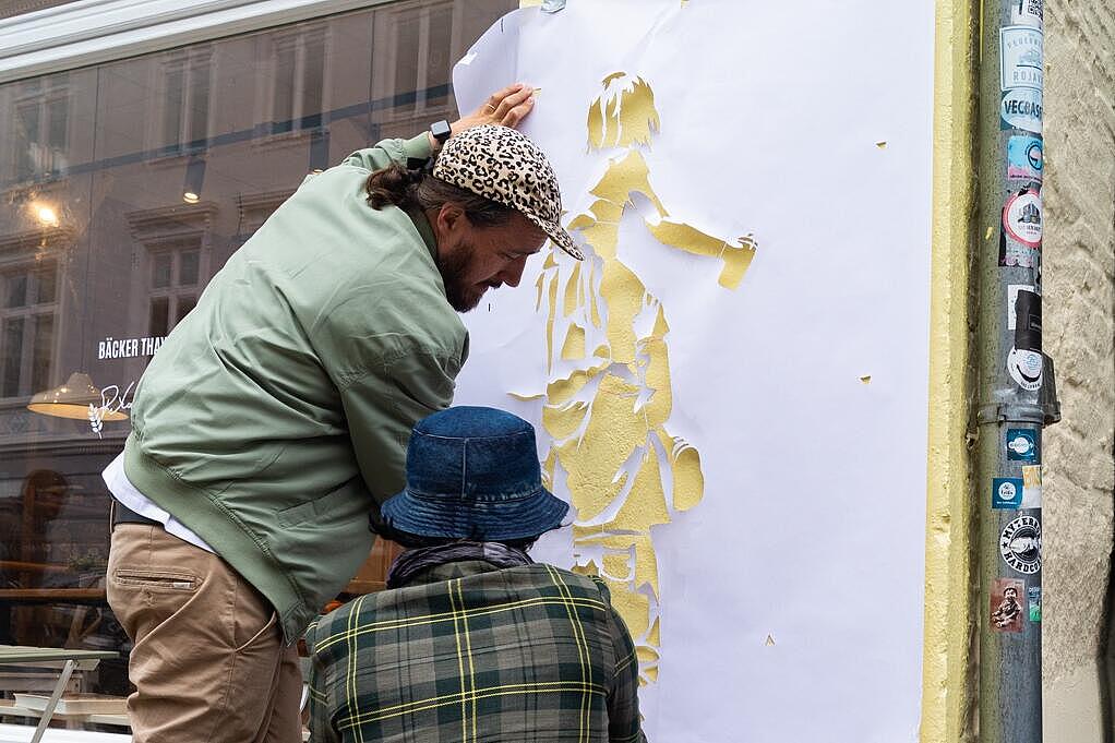 Zwei Personen befestigen eine Papierschablone an der Außenwand einer Bäckerei in der Flensburger Innenstadt.