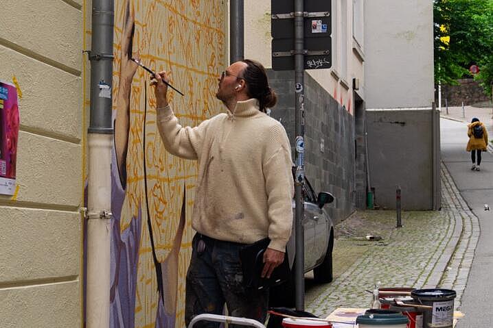 Ein Mann malt mit einem Pinsel ein Gemälde an eine Hauswand. Auf einem Tisch neben ihm stehen mehrere Farbeimer.