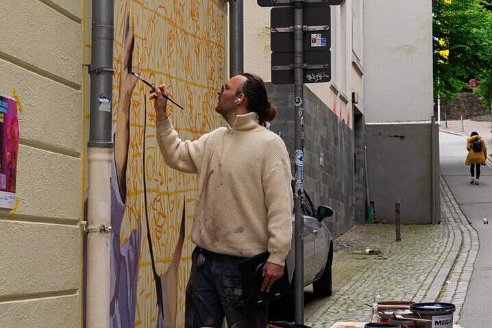 Ein Mann malt mit einem Pinsel ein Gemälde an eine Hauswand. Auf einem Tisch neben ihm stehen mehrere Farbeimer.