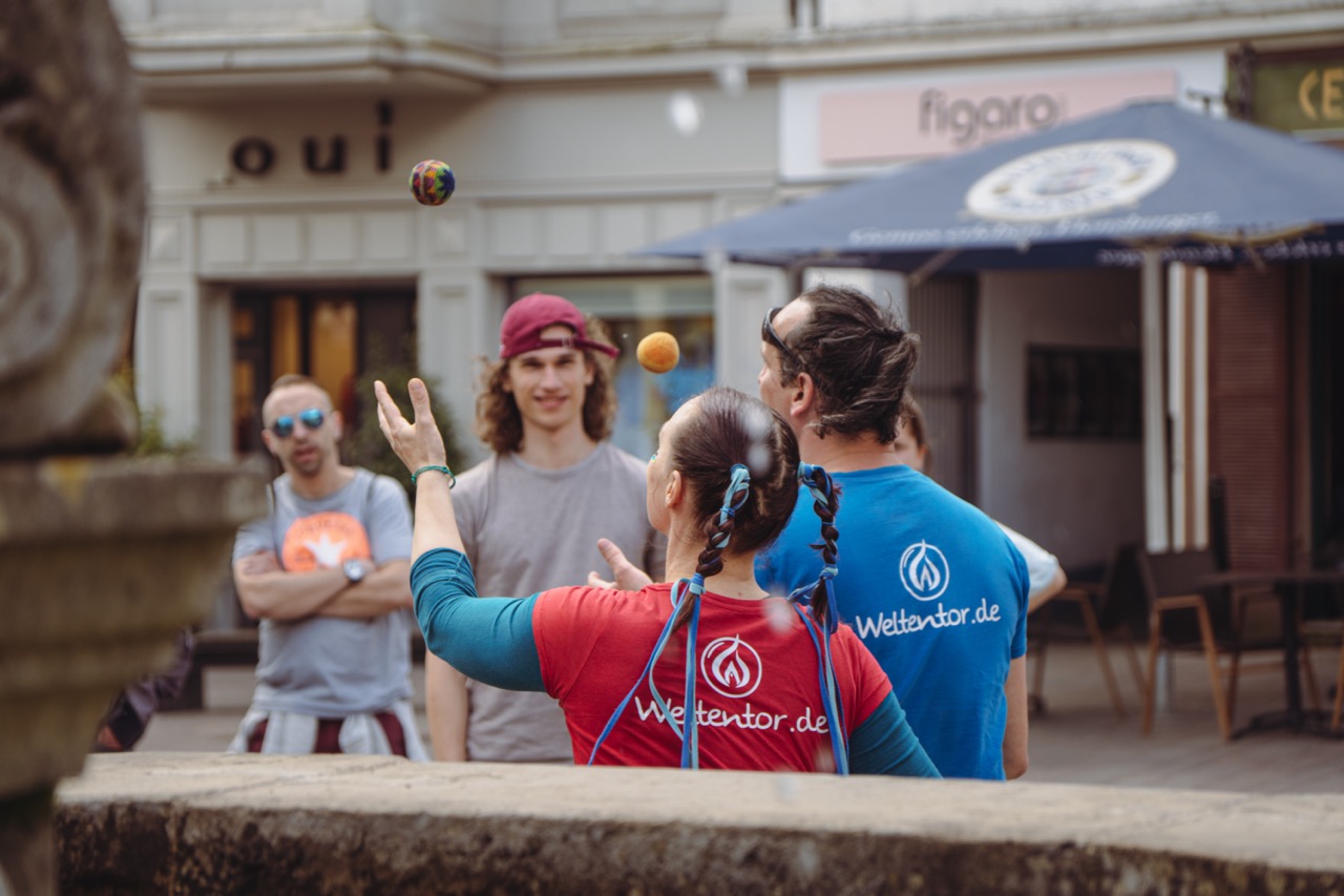 Am Neptunbrunnen in Flensburg stehen zwei Personen in T-Shirts mit der Aufschrift Weltentor.de und werfen jeweils einen Jonglierball in die Luft. 