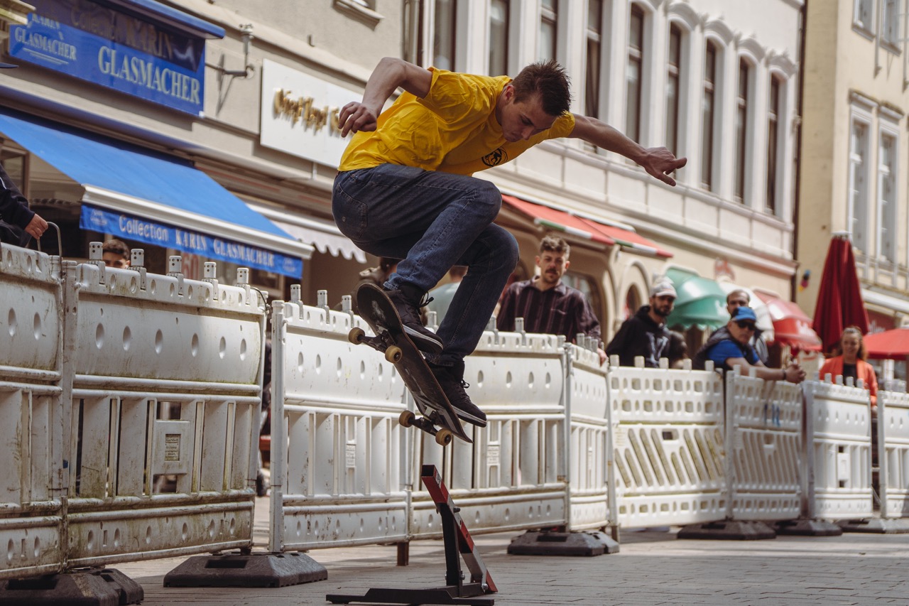 In der Flensburger Fußgängerzone springt ein Mann mit einem Skateboard in die Luft. Hinter ihm ist ein Schrankenzaun aufgebaut, an dem Besuchende stehen und zuschauen.