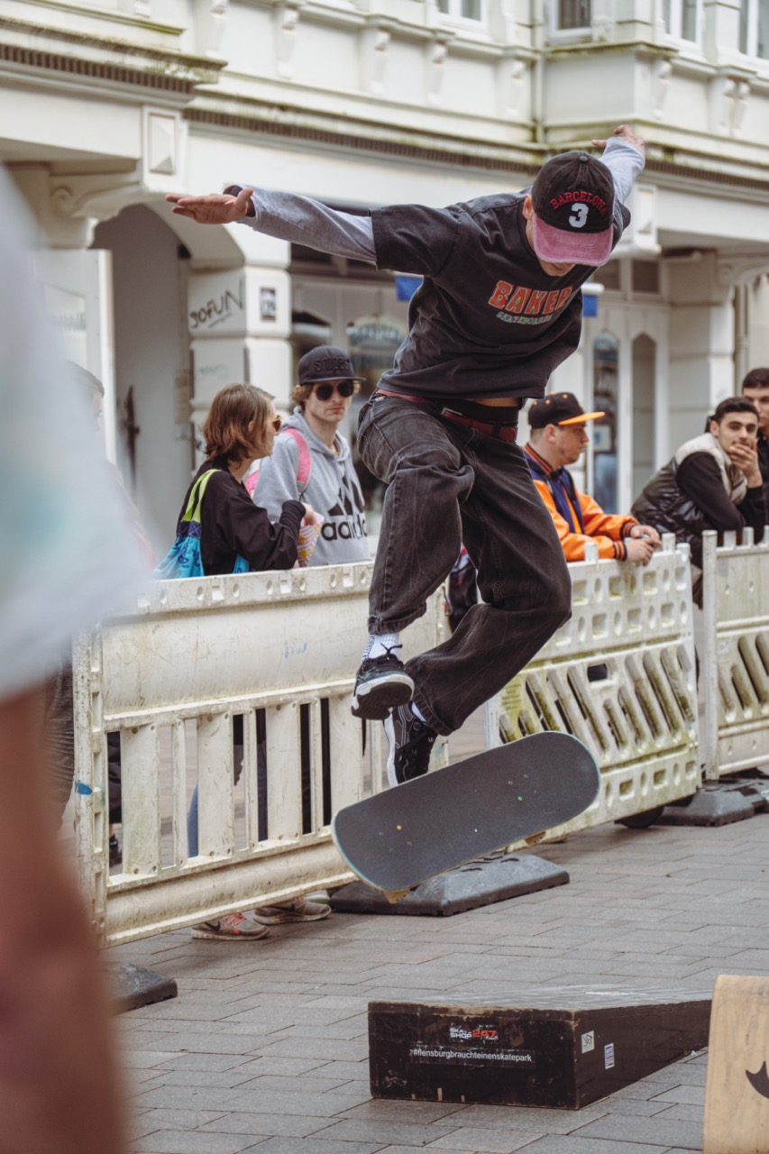 In der Flensburger Fußgängerzone springt ein Mann an einer Rampe mit einem Skateboard in die Luft. Hinter ihm ist ein Schrankenzaun aufgebaut, an dem Besuchende stehen und zuschauen.