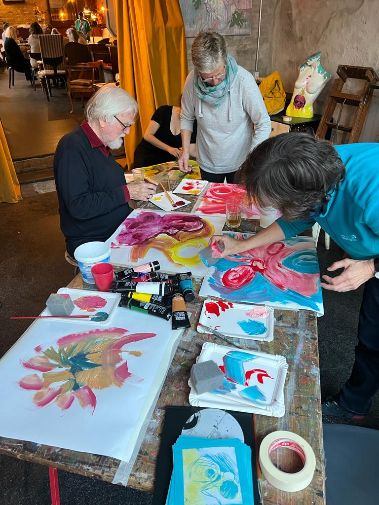 Besuchende des Kunst-Events zur Menschenwürde malen an einem Tisch farbenfrohe Bilder mithilfe von Pinseln und Schwämmen.