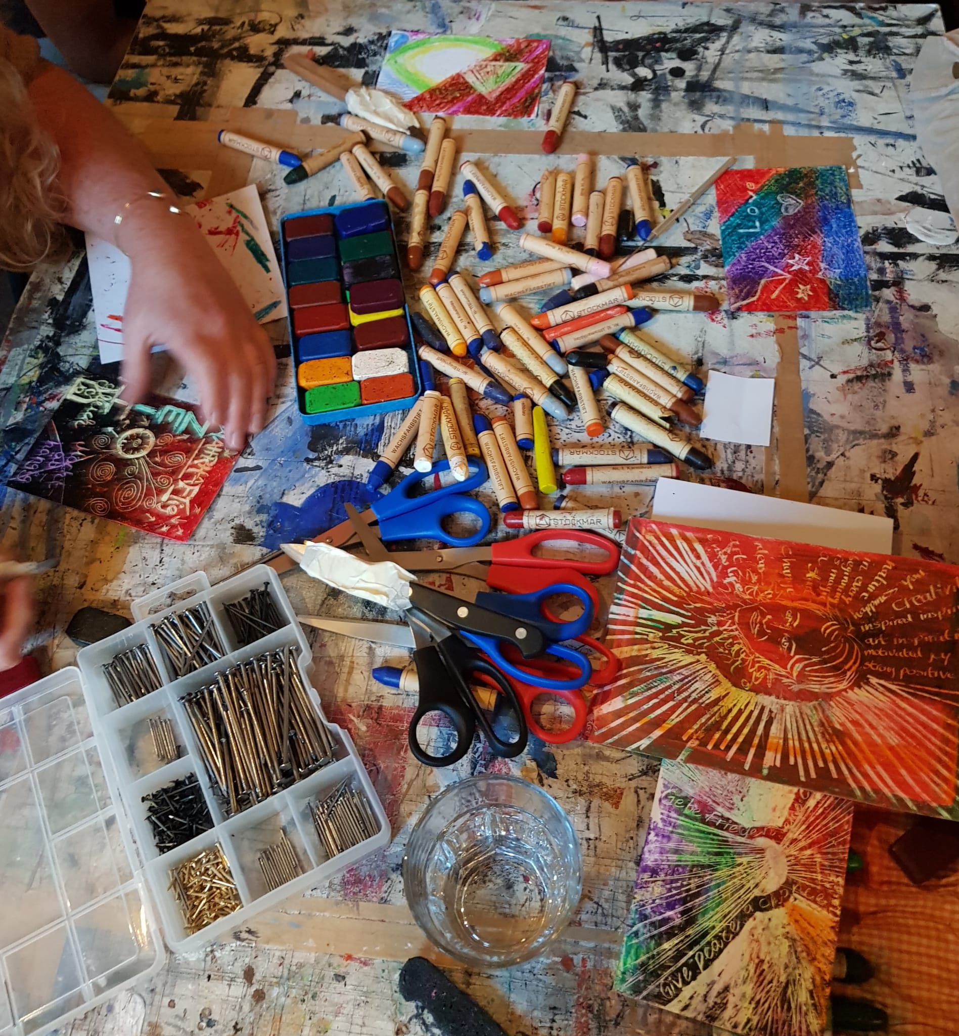 Auf einem Tisch liegen viele bunte Wachfarben, Scheren, ein Kasten mit Nägeln unterschiedlicher Größen sowie selbst gemalte Wachsbilder.