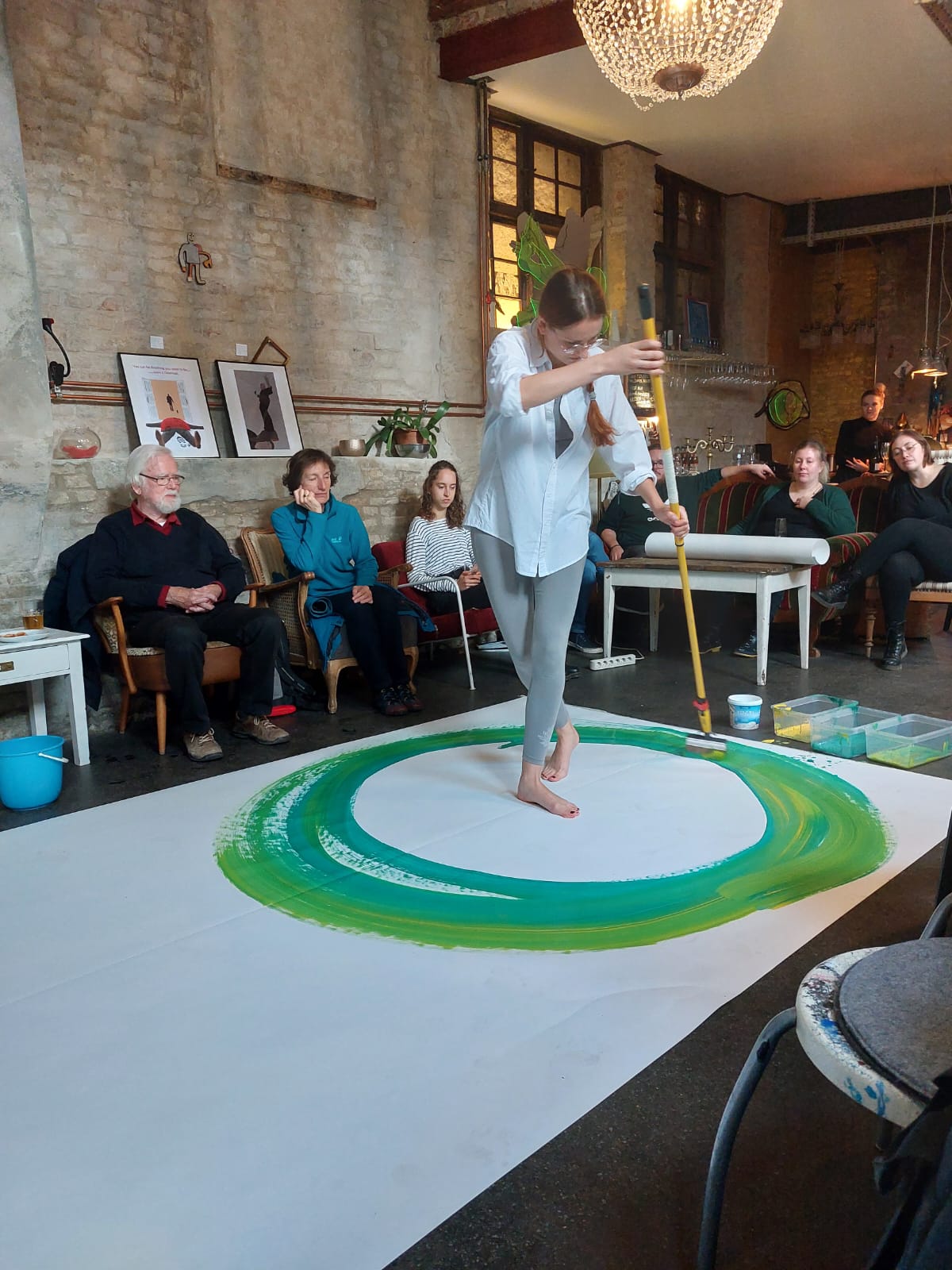 Eine Frau steht auf einem übergroßen weißen Papier und malt mit unterschiedlichen Grüntönen einen Kreis um sich herum. Auf Sesseln und einem Sofa sitzen Besuchende des Kunstevents zur Menschenwürde und schauen zu.
