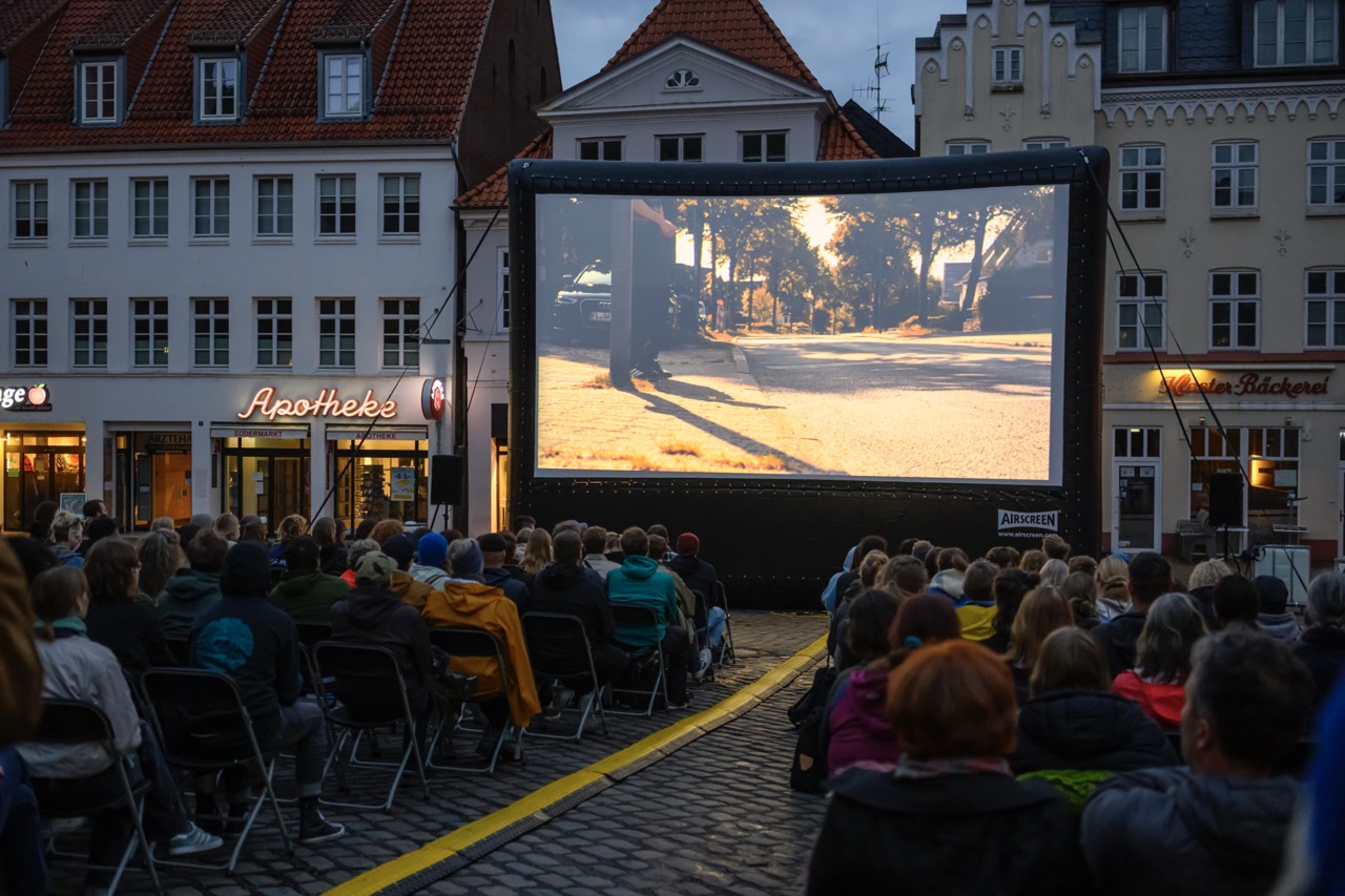 Menschen sitzen auf Klappstühlen vor einer großen Leinwand auf dem Südermarkt in Flensburg.