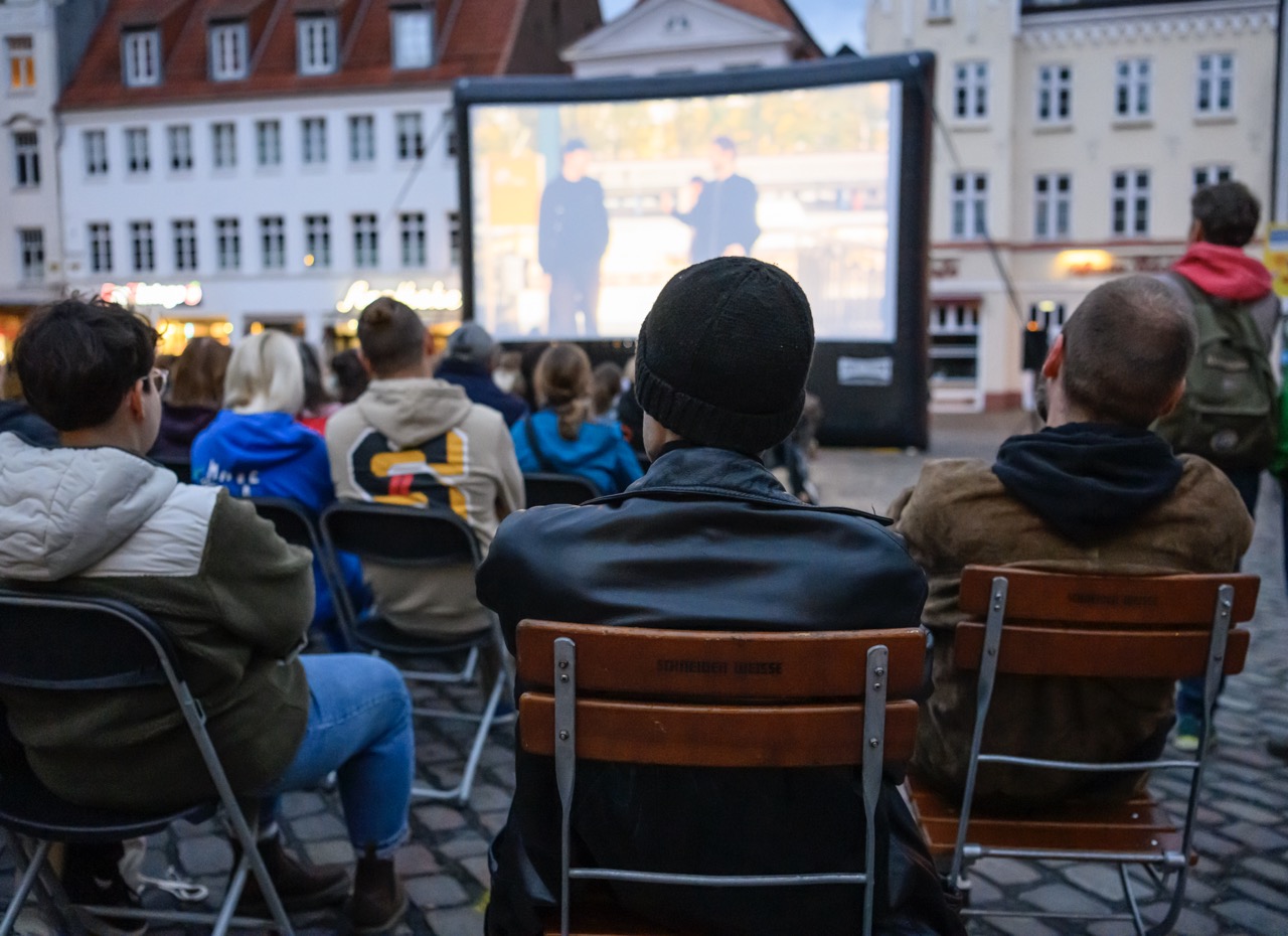 Menschen sitzen auf Klappstühlen vor einer großen Leinwand auf dem Südermarkt in Flensburg.