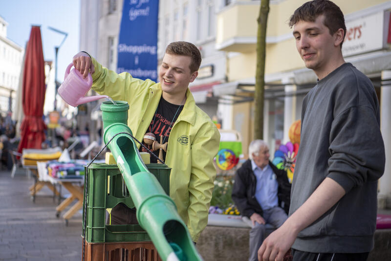 Eine Person gießt Wasser in eine selbst gebaute Wasserbahn in der Flensburger Fußgängerzone. 