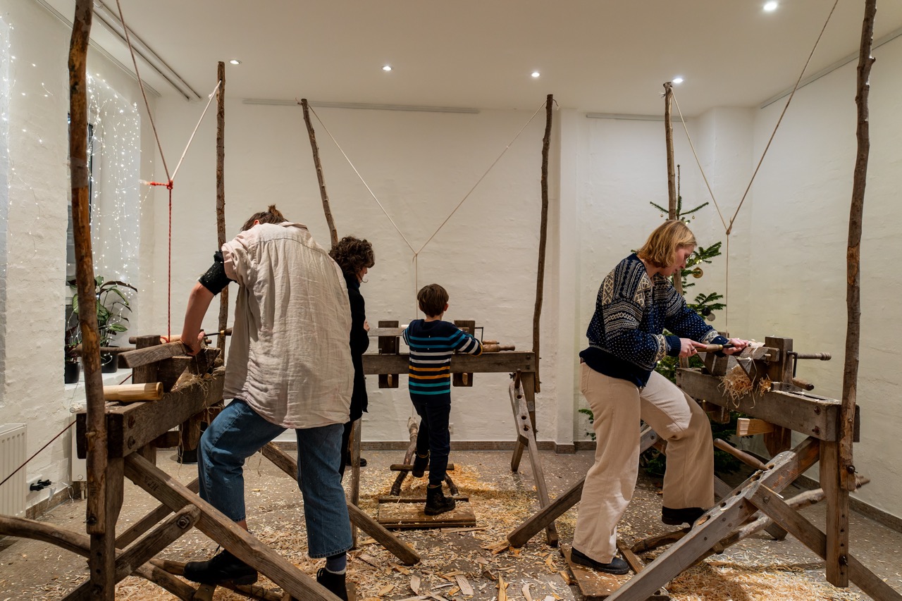 Drei Erwachsene und ein Kind arbeiten an Werkbänken im schönHIER! in Flensburg. In der Ecke des Raums steht ein geschmückter Weihnachtsbaum.