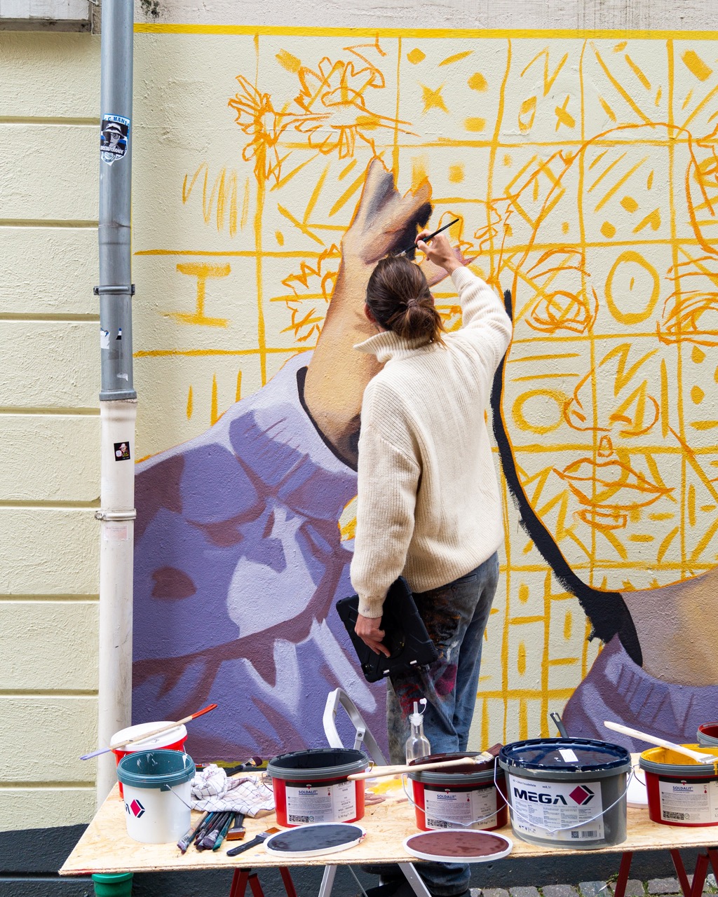 Ein Mann malt mit einem Pinsel ein Gemälde an eine Hauswand. Auf einem Tisch neben ihm stehen mehrere Farbeimer sowie Malutensilien.