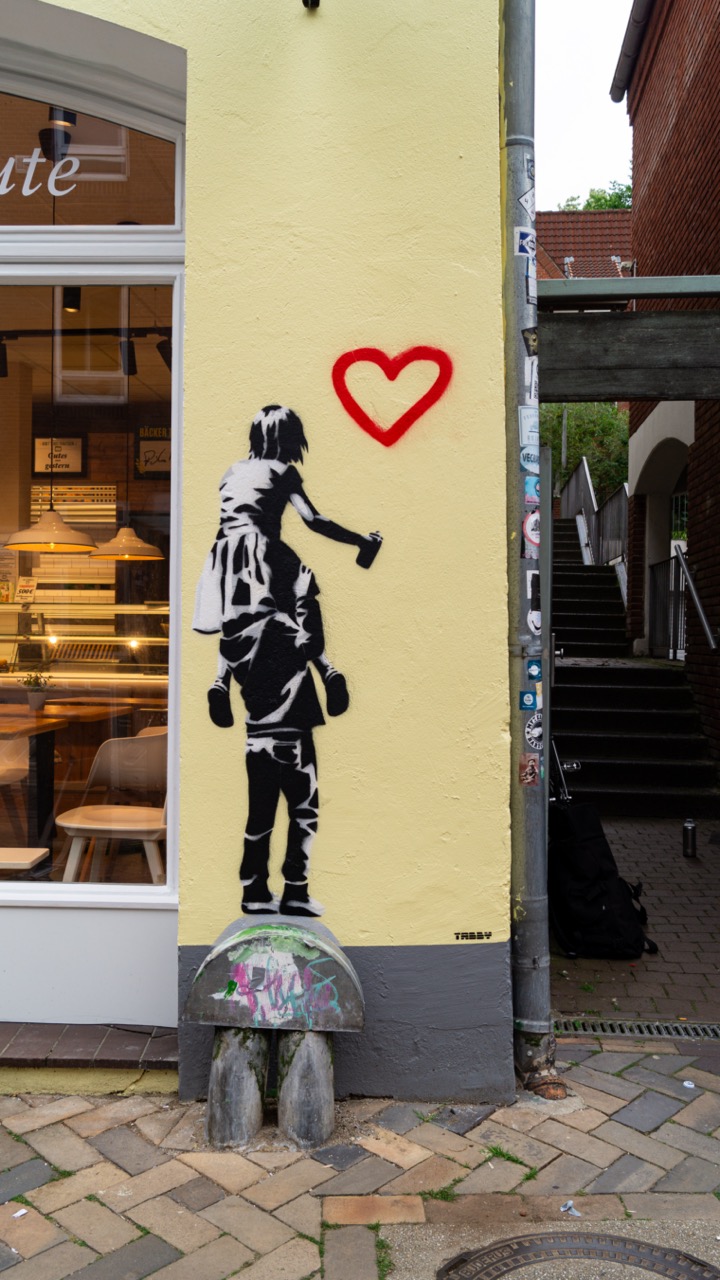 Streetart auf der Außenwand einer Bäckerei in der Flensburger Innenstadt. Sie zeigt eine Person mit einer Spraydose in der Hand, die auf den Schultern einer anderen Person sitzt. Über den beiden ist der Umriss eines roten Herzens gesprayt.