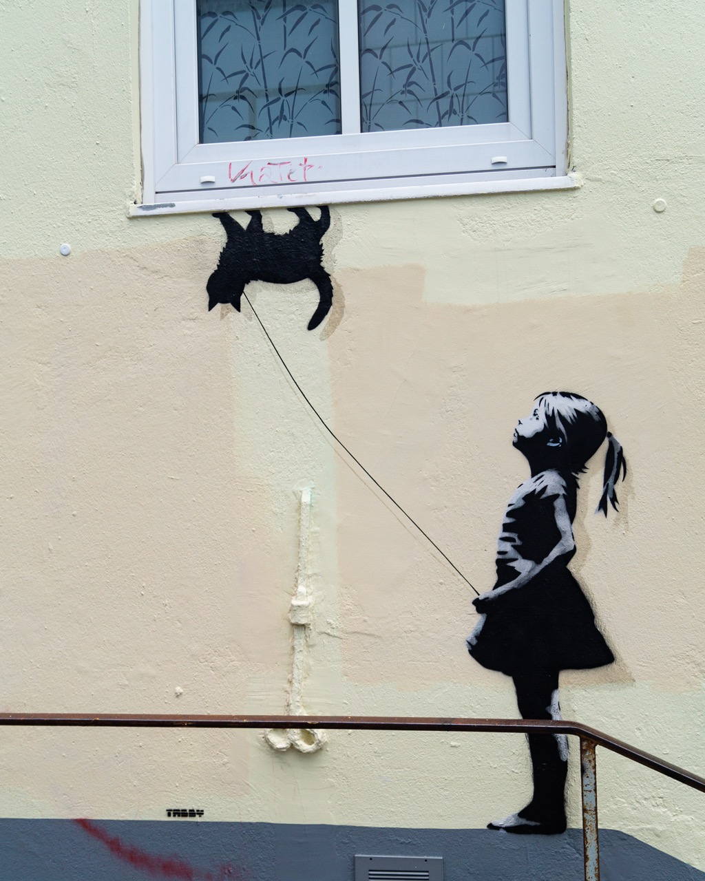 Streetart auf einer Außenwand in der Flensburger Innenstadt. Sie zeigt ein Mädchen, das eine Katze an der Leine führt und zu ihr hoch schaut. Die Katze steht Kopfüber unter einem Fenster.