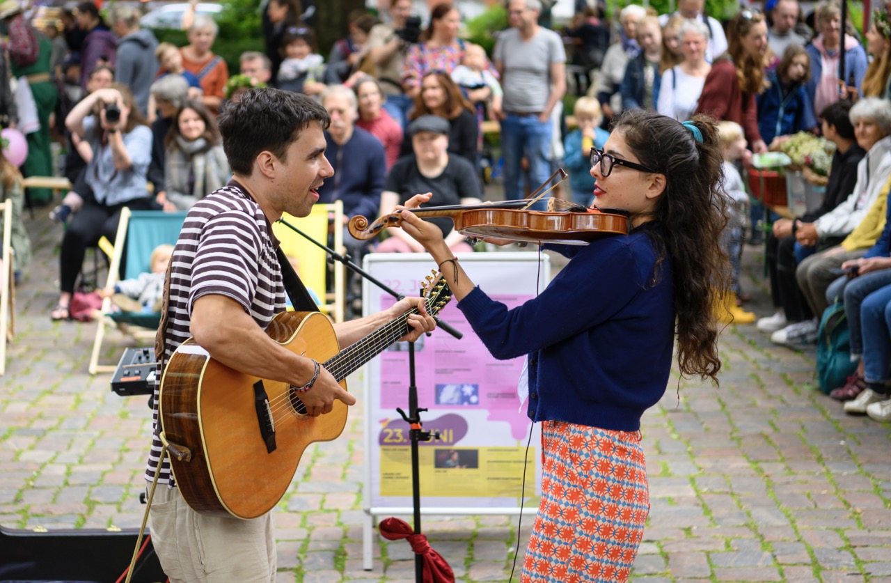 Ein Gittarist und eine Violonistin spielen vor Publikum unter freiem Himmel.