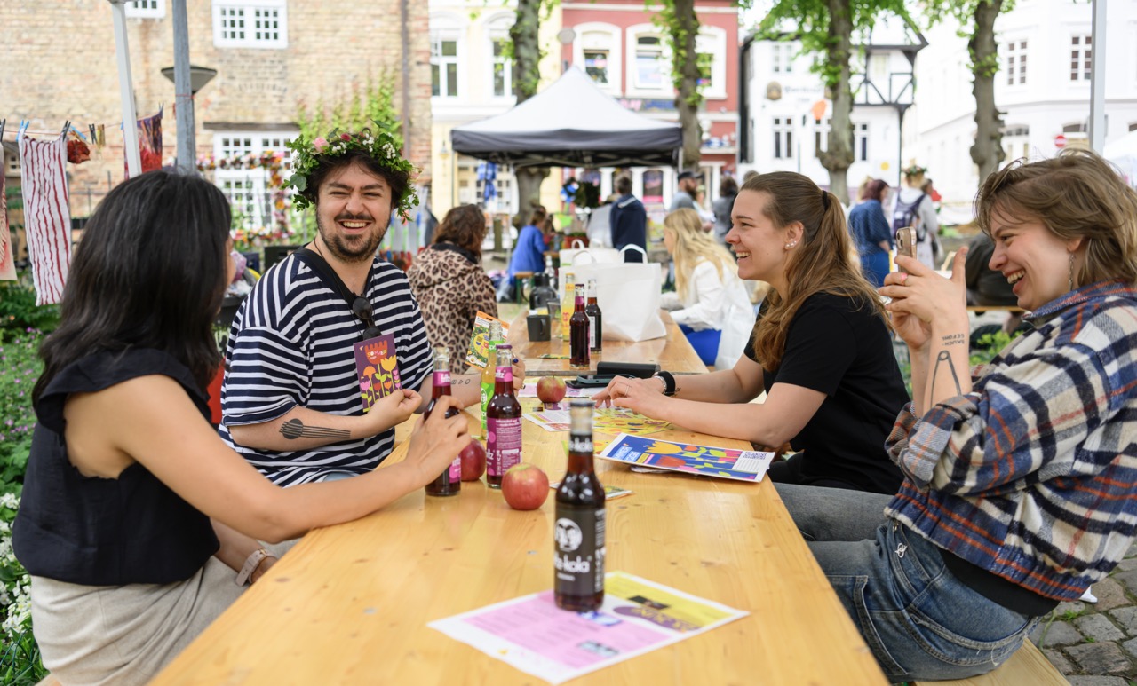 Lachende Gäste der Veranstaltung Sommer Hygge sitzen an Biertischen auf dem Hof der Marienkirche Flensburg.