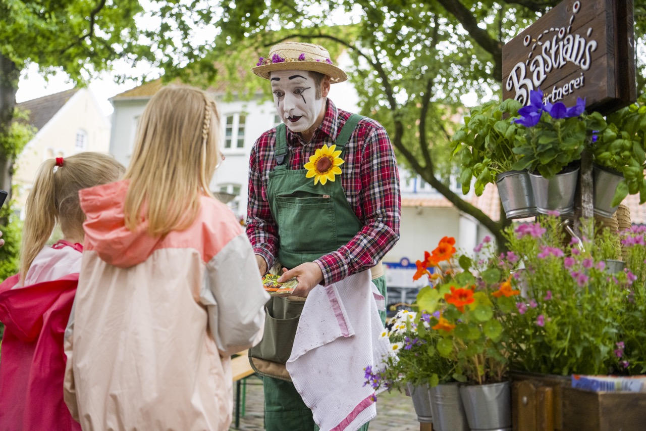 Eine wie ein Clown geschminkte Person sowie zwei Kinder vor einem Blumenstand mit der Beschriftung Bastians Gärtnerei.